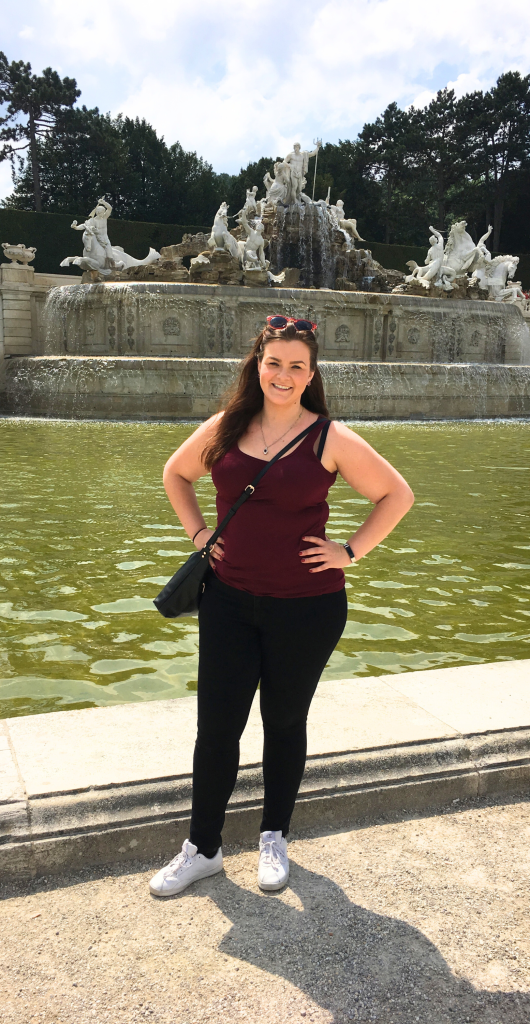 Where to Stay in Vienna, Austria: My Favorite Areas + Best Budget Hotels 7 Young woman smiles by a stunning ornamental fountain in a vibrant garden setting in Vienna Austria.