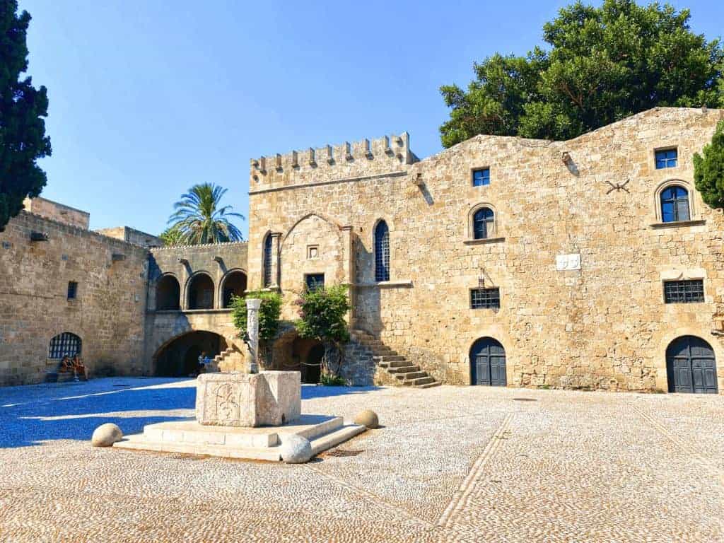 3-Day Rhodes Itinerary: How to Spend Time on Greece's Medieval Island 19 Medieval stone walls and tower at Argyrokastrou Square in the Old Town of Rhodes, Greece, framed by trees and historic buildings in a sunlit open plaza.