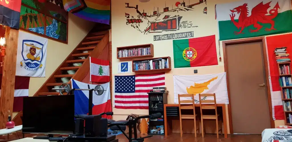 Are Hostels Safe? Honest Experiences from a Decade of Travel + 10 Takeaways 10 Common area in the Loft Hostel of Budapest, Hungary, features numerous flags from different countries, including the USA, Portugal, Wales, and Cyprus as well as workout equipment, a desk, tv, and bookshelves.