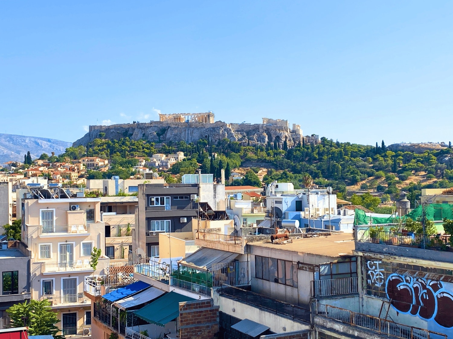 View from Downtown Athens Lofts in Greece, showing the ancient Acropolis with the Parthenon perched atop its rocky hill rising above a cluster of modern buildings and apartment rooftops under a clear blue sky.
