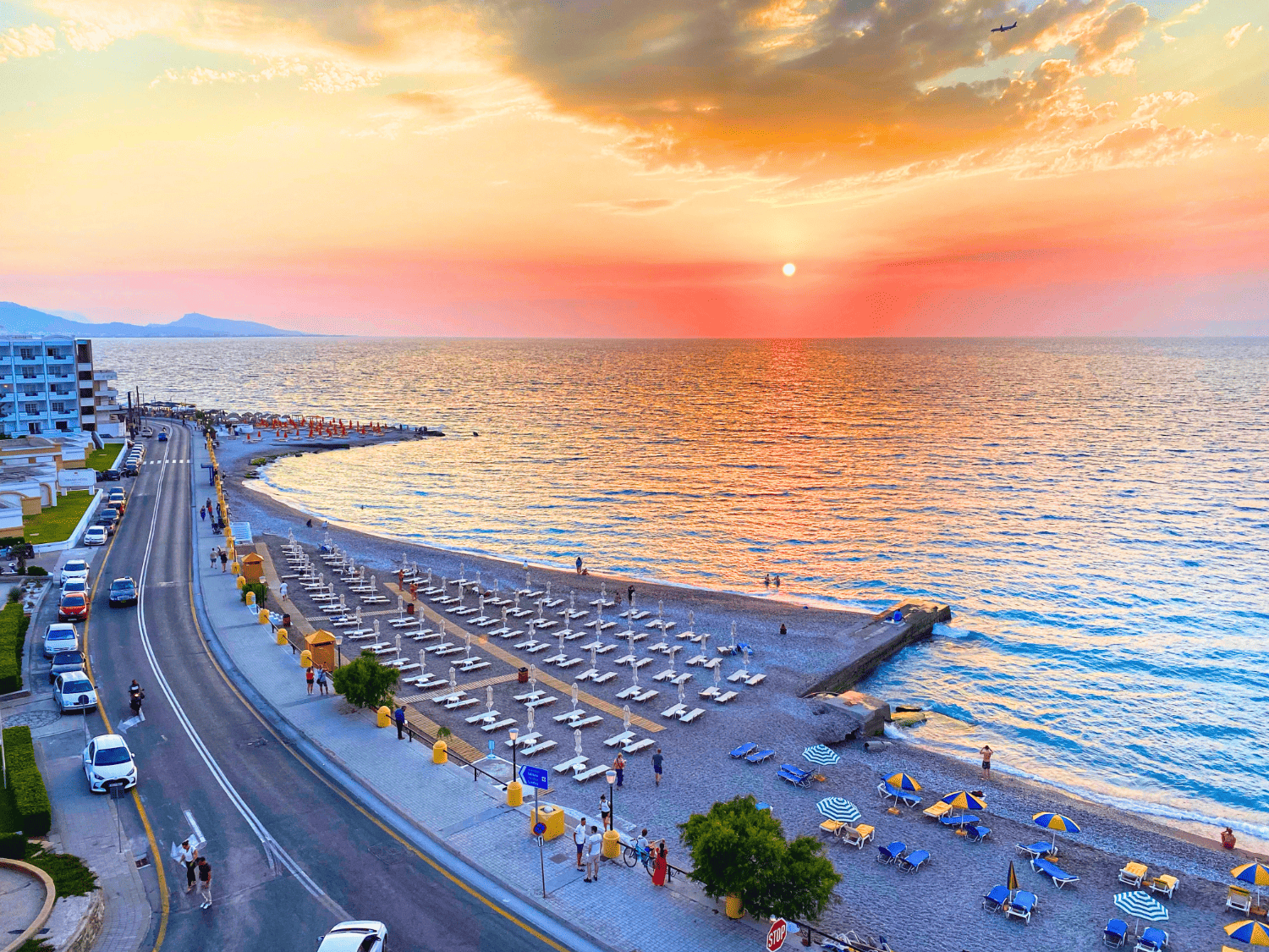 View from Mitsis La Vita Beach Hotel of the sunset over Elli Beach in Rhodes, Greece, showing the golden sun dipping toward the horizon and casting warm orange and pink light across the calm sea and sandy shore with silhouettes of distant rocks or headlands.