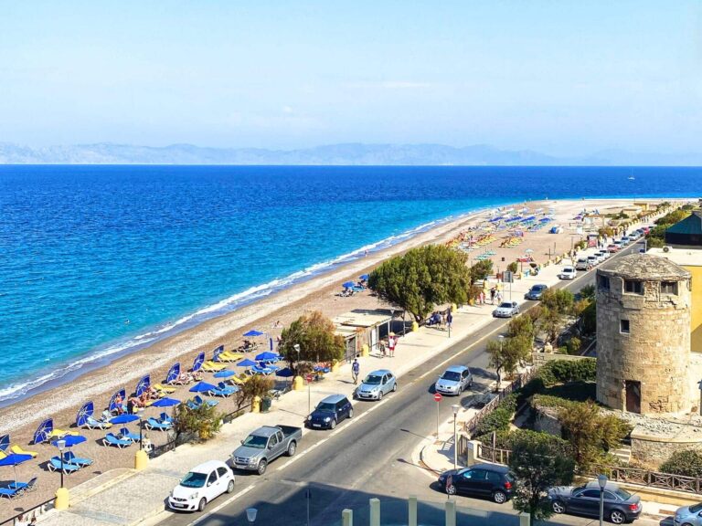 View from Mitsis La Vita Beach Hotel of Elli Beach in Rhodes Town, Greece, featuring a long curve of pebbly shoreline with rows of colorful beach umbrellas and sun loungers beside the clear turquoise Aegean Sea.