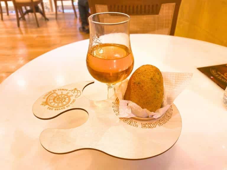 A pastel de bacalhau, also known as a codfish cake or croquette, is served golden and crispy from Casa Portuguesa do Pastel de Bacalhau in Lisbon, Portugal. The savory treat is presented in a paper wrapper alongside a glass of port wine.