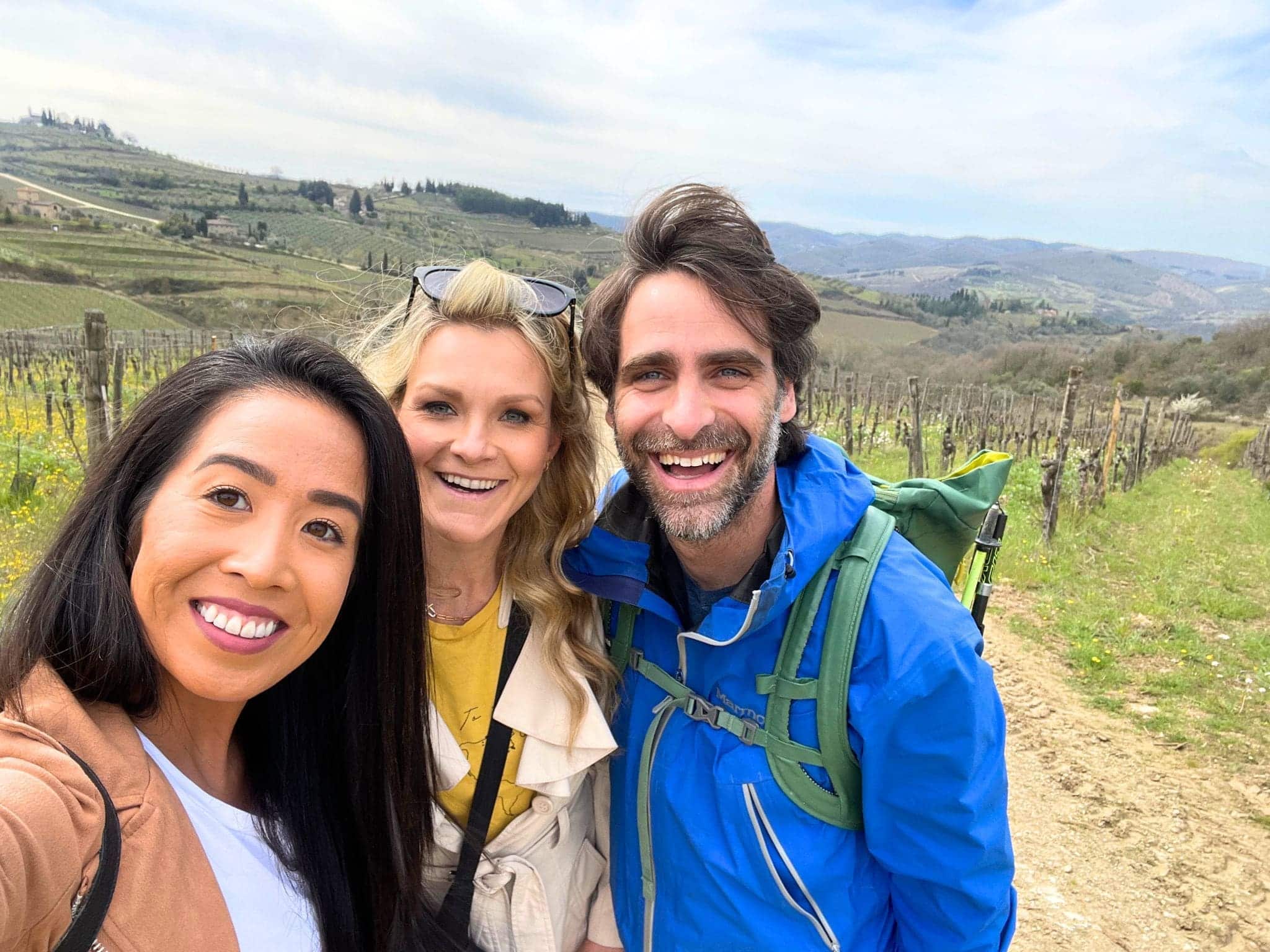 Michelle L. Jensen (Travel HerStory) and two friends pose in sunlit vineyard in Tuscany while on a wine tasting tour, smiling and enjoying the scenic countryside. Rows of grapevines stretch into the distance under a clear sky in Italy.