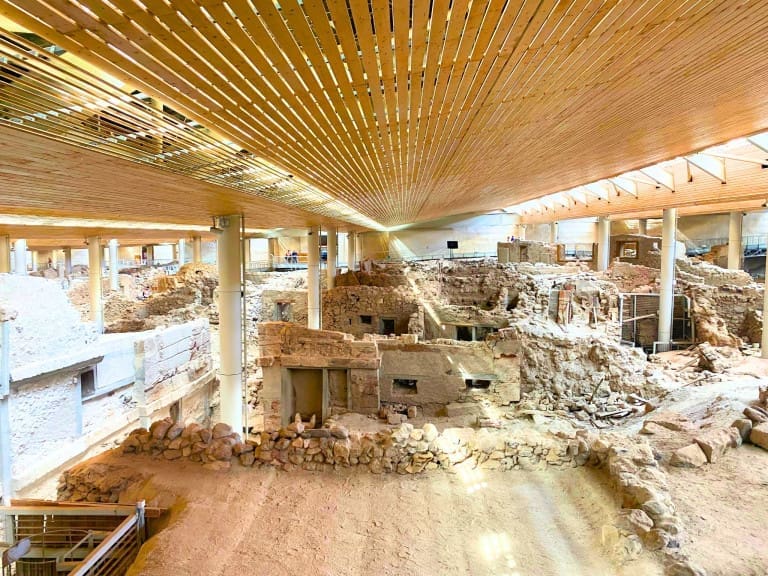Interior view of the Akrotiri archaeological site on Santorini, Greece, showing excavated ancient stone buildings and streets preserved under a modern protective roof with wooden beams and support columns above.