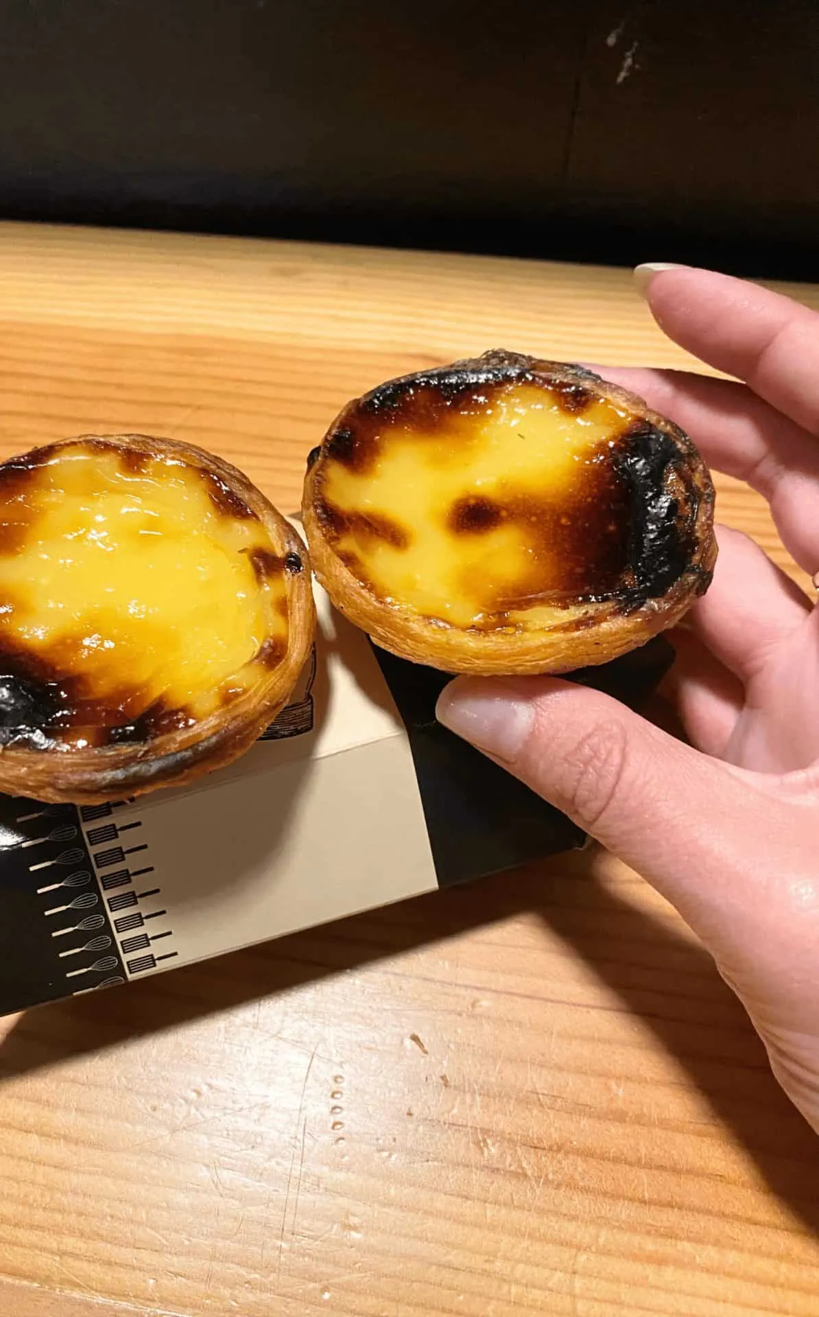 Home 36 Hand holding freshly baked pastéis de nata with golden custard centers and flaky crusts on top of a box from Manteigaria at the Time Out Market in Lisbon, Portugal.