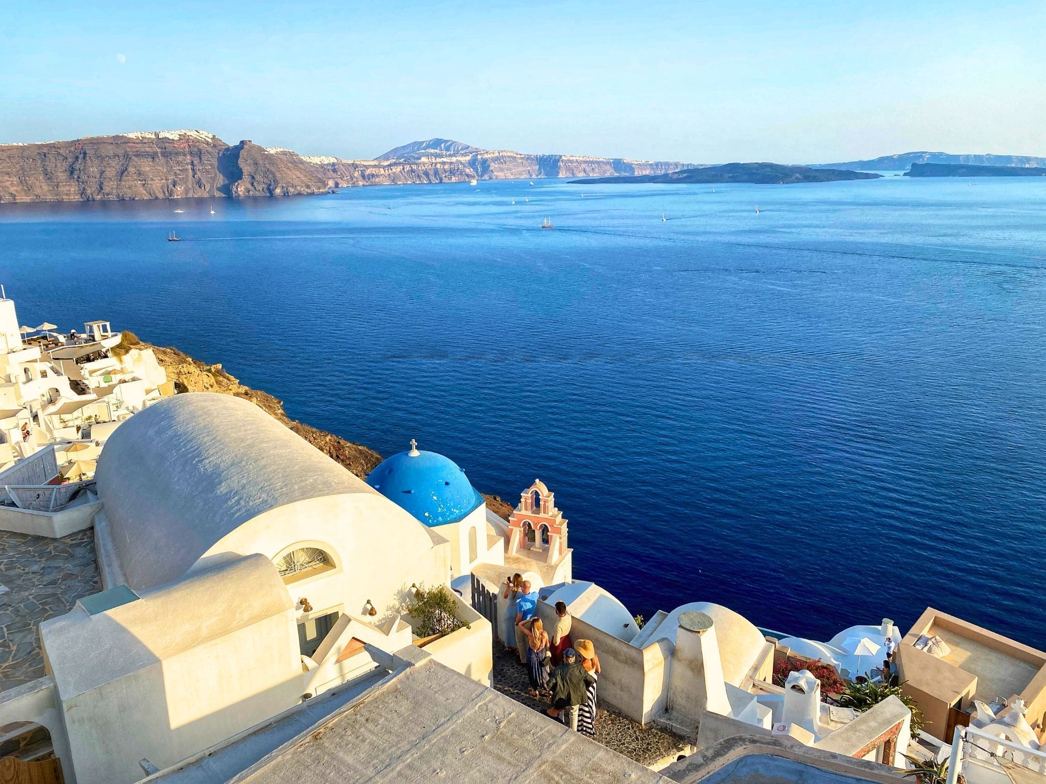 Greece 43 Golden light of sunset hits the Santorini caldera as seen from the terrace of Pelekanos Restaurant in the village of Oia, Greece, featuring whitewashed buildings and blue-domed churches cascading down the cliffside toward the deep blue Aegean Sea.