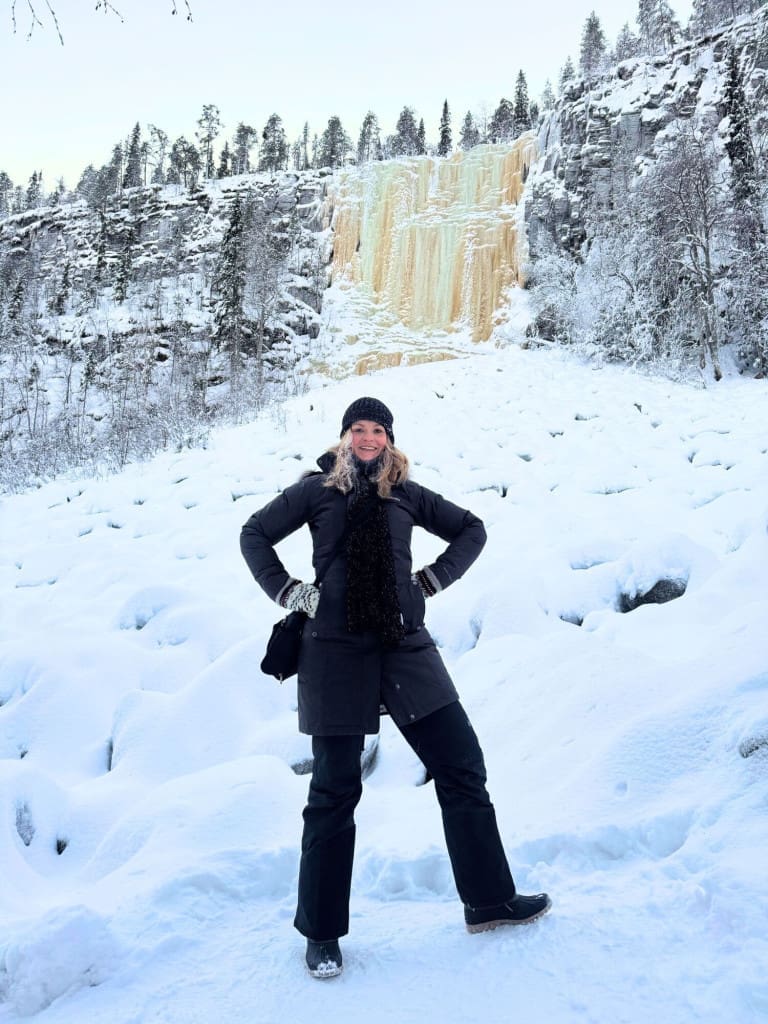 30 of the Best Things to Do in Rovaniemi, Finland, This Winter 84 Woman standing in Finnish snow at frozen waterfall in scenic winter landscape while holding a Travelon crossbody bag.