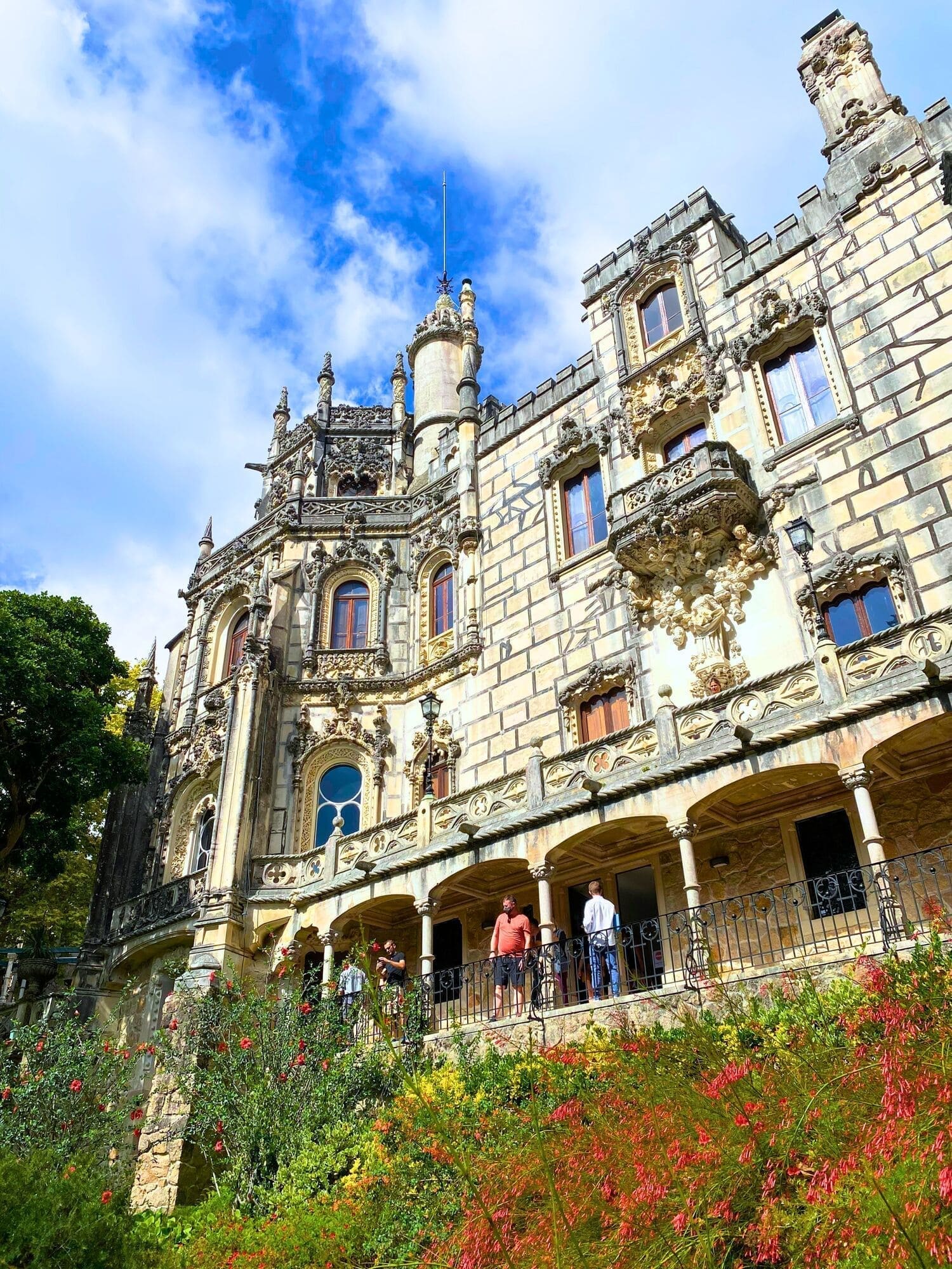 Portugal 17 Upward view of the exterior of Quinta da Regaleira in Sintra Portugal showing the ornate Romantic‑era manor house with intricate Gothic and Manueline architectural details set against lush gardens and trees. The historic estate’s decorative towers and stone façades rise above the landscaped grounds of this UNESCO World Heritage Site as people stand on the balcony.