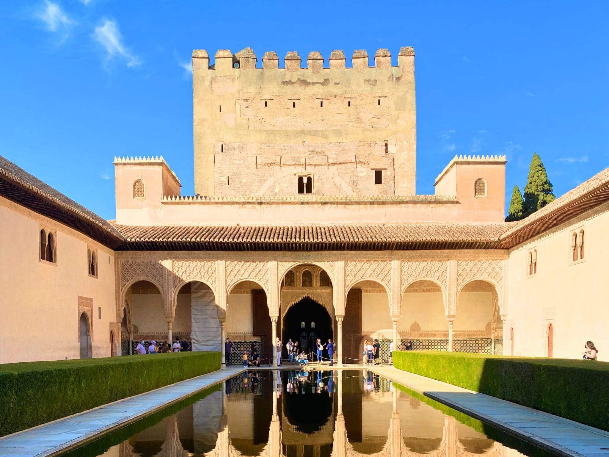 Court of the Myrtles within the Nasrid Palaces at the Alhambra complex in Granada, Spain, looking north towards the Comares Tower, featuring a long reflective pool lined with myrtle bushes and bordered by Moorish arches and intricate stucco walls. The courtyard exemplifies the symmetry and elegance of Islamic palace architecture.