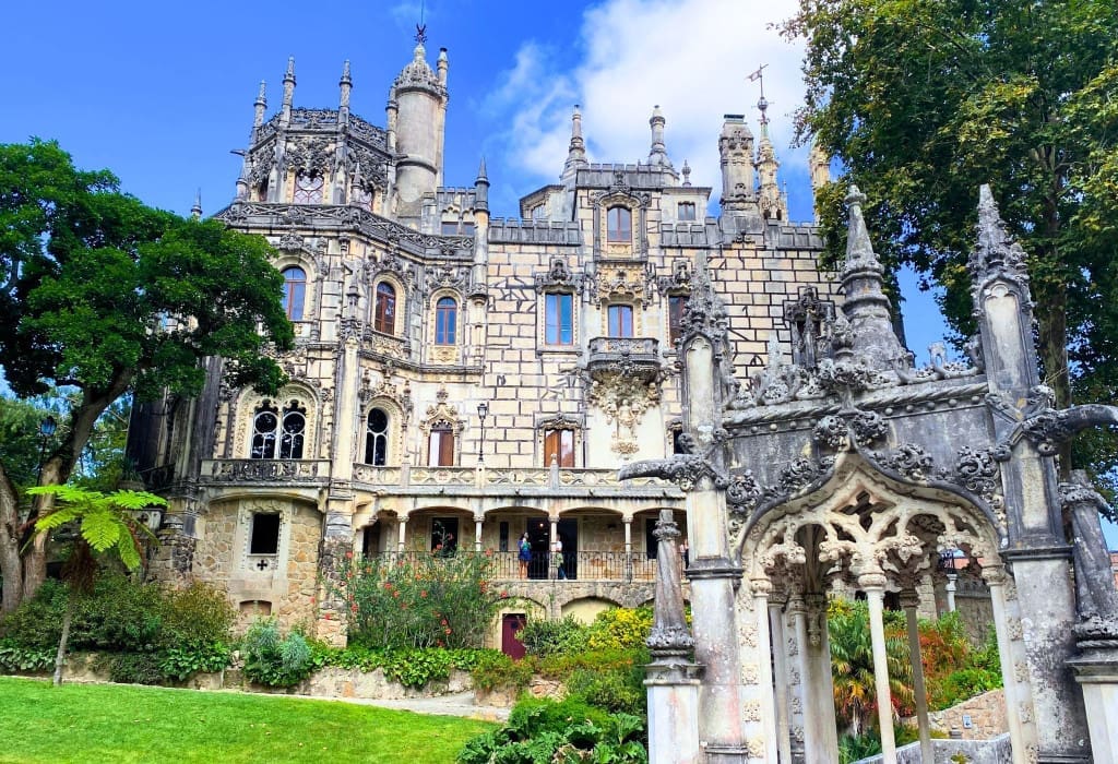 The Top Things to Do in Sintra: 5 Stunning Palaces You Need to See 12 Front facade of Quinta da Regaleira palace in Sintra, Portugal, viewed from the front lawn with an ornate carved stone pinnacle gateway in the foreground. The neo-Manueline tower and turrets rise behind visitors strolling the covered veranda, framed by garden trees under a blue sky.