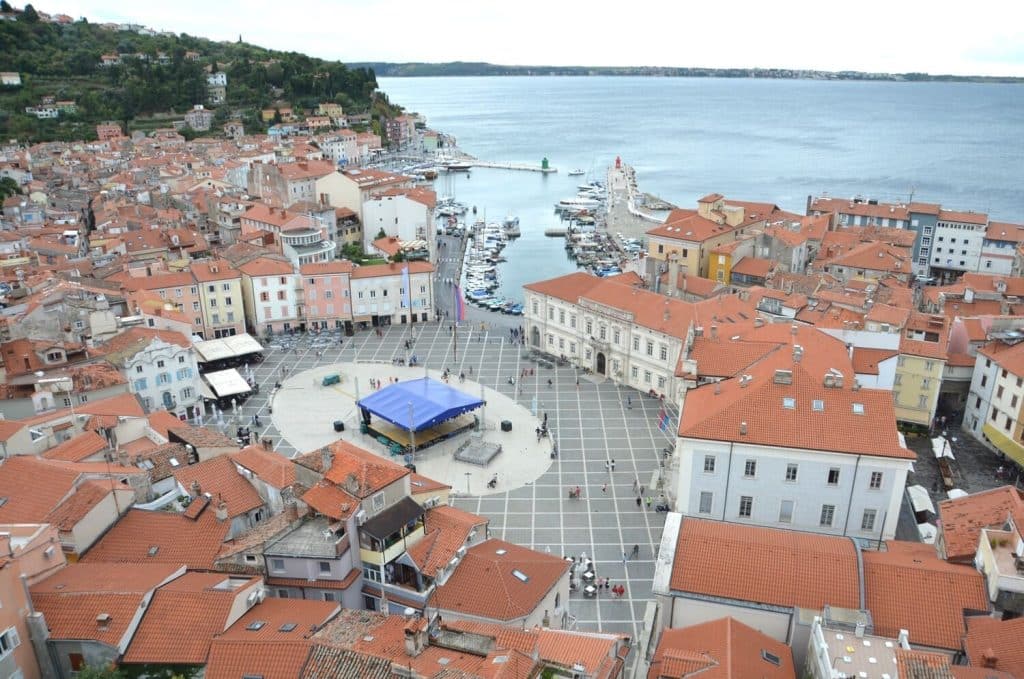 18 of the Best Things to Do in Slovenia: Your Ultimate Travel Guide 1 An aerial view of the historic coastal town of Piran, Slovenia, focusing on the main Tartini Square. Surrounded by red-roofed buildings and set against the blue Adriatic Sea, the square features a statue of Giuseppe Tartini and a stage setup for an event.