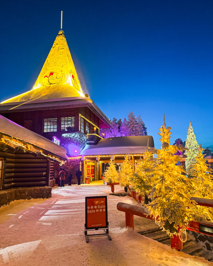 The Best Hotels in Rovaniemi, Finland: Comparing My Glass Igloo Stay with the City Center 60 Santas Village in Rovaniemi, illuminated at night, with snow-covered trees and festive lights.