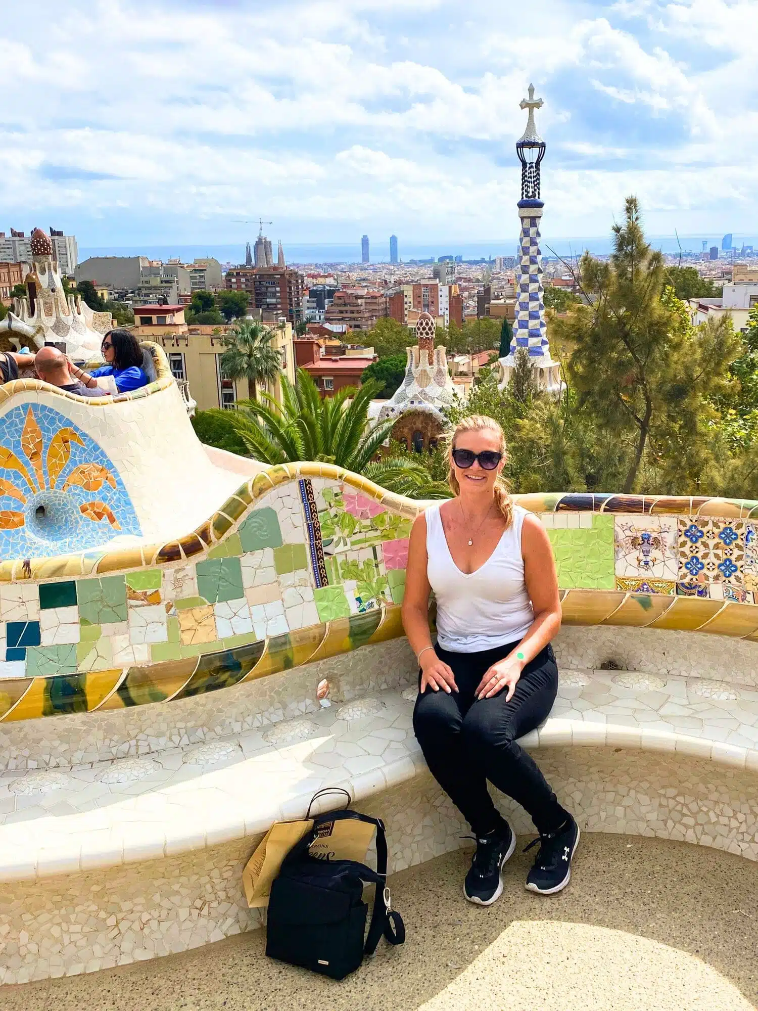 Home 35 Michelle L. Jensen (Travel HerStory) seated on the iconic serpentine bench at Park Güell in Barcelona, Spain, designed by Josep M. Jujol. The bench, covered in vibrant tile-shard mosaic, winds along the terrace edge and doubles as a balustrade overlooking the park’s architectural landscape with the city in the background.