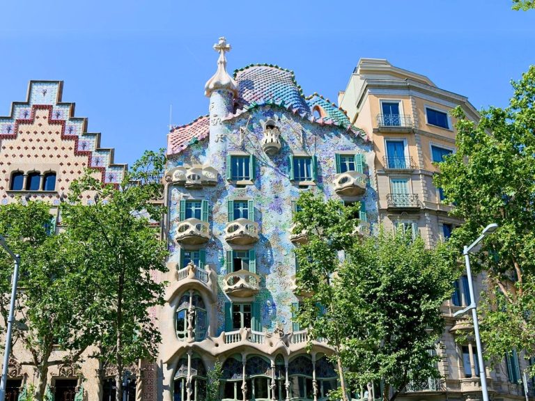 Front facade of Casa Batlló in Barcelona, Spain, a masterpiece by Antoni Gaudí, with its colorful mosaic surface, flowing stone balconies, and organic window shapes. The undulating design and skeletal features give the building its iconic, otherworldly appearance, often likened to the surface of a dragon or sea creature.