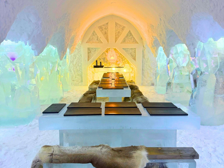 Mining-themed architectural ice sculptures inside the Arctic SnowHotel & Glass Igloos Ice restaurant in Sinettä, Finland, featuring a lantern, tables of ice, and fur hides glowing warmly in the frozen space.