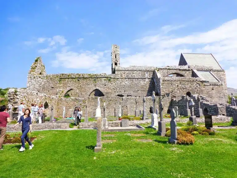 Tourists exploring the ancient stone ruins of a Benedictine Abbey and a historic graveyard in the Irish countryside under a blue sky.