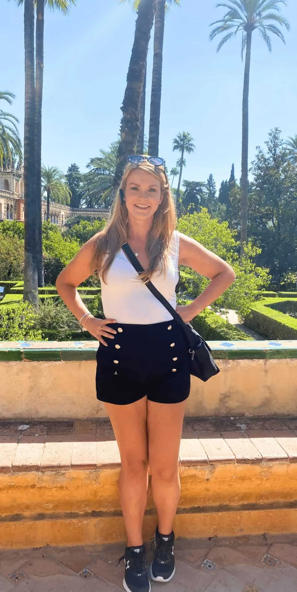 Home 33 Michelle L. Jensen (Travel HerStory) poses in the sunlit garden of the Royal Alcázar of Seville, Spain, surrounded by trimmed hedges, palm trees, and ornate Moorish architecture while wearing an audioguide, Travelon purse, and sunglasses.