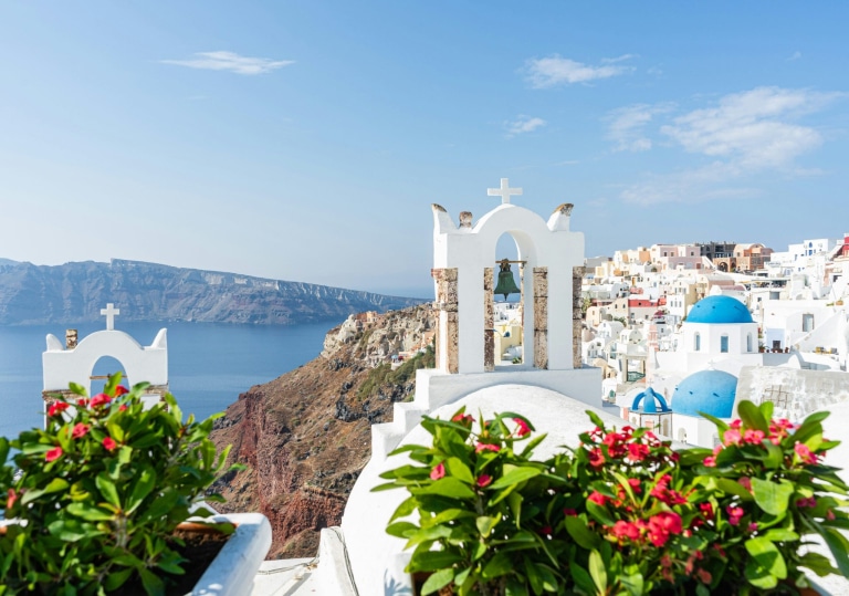 Over a church with a bell tower on the caldera in Santorini, Greece, with whitewashed buildings and blue-domed churches clinging to the cliffside with the view extending across the Aegean Sea.
