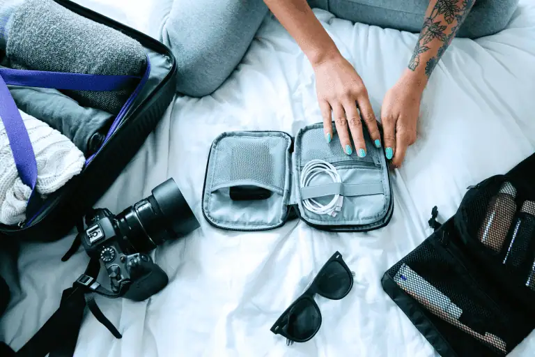 Woman packing for trip with a camera, sunglasses, and packing cubes for organizing essentials on a bed next to a suitcase.