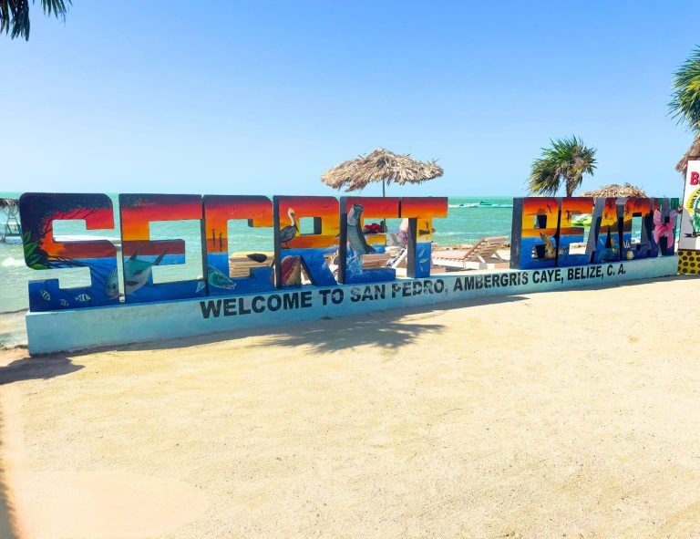 Colourful large letter Secret Beach sign on the sandy shore at Ambergris Caye, Belize, with tropical marine artwork painted across each letter, reading Welcome to San Pedro, Ambergris Caye, Belize C.A., with turquoise Caribbean water, a palapa umbrella, and palm trees visible behind