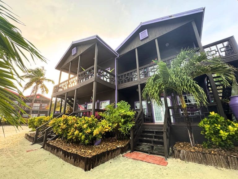 Where's the Best Place to Stay in Ambergris Caye, Belize? Luxury Resort or Budget Hostel 6 Exterior view of a Pool View Villa at Xtan Ha Resort on Ambergris Caye, Belize, showing two-storey dark timber beachfront villas with wrap-around balconies, purple accent details, wooden staircases, hammocks on the lower level, and lush tropical garden landscaping on white sandy beach at sunset.