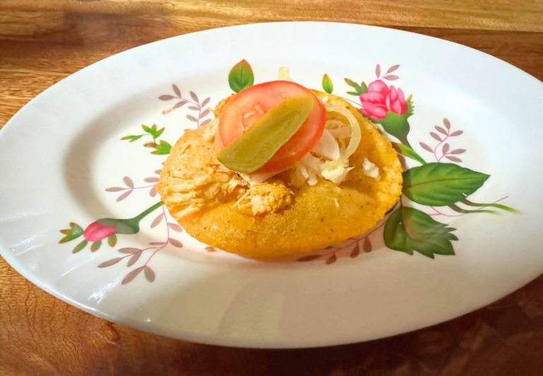 A traditional Belizean salbute, a golden fried masa tortilla topped with shredded chicken, sliced tomato, pickled jalapeño, and white onion, served on a white floral-patterned plate at Briana’s Tropical Taste in San Pedro, Ambergris Caye, Belize on a wooden table.