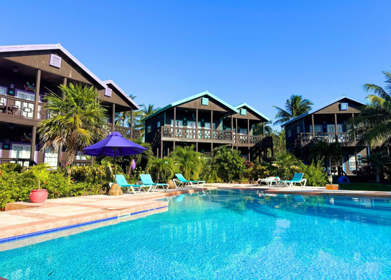 Turquoise swimming pool at Xtan Ha Resort on Ambergris Caye, Belize, surrounded by teal sun loungers, a large purple parasol, lush tropical palm trees and gardens, with two-storey dark timber villas with wrap-around balconies and colourful accent details visible behind, under a vivid blue sky.