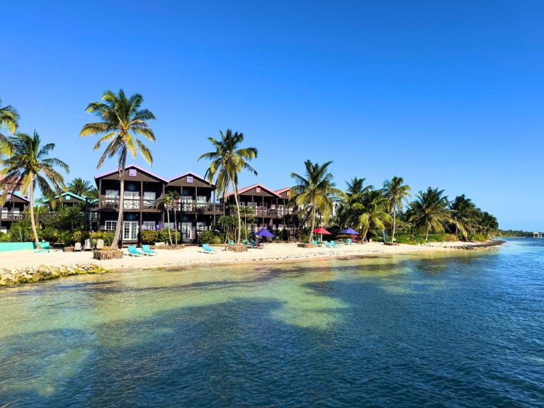 Panoramic waterfront view of Xtan Ha Resort on Ambergris Caye, Belize, from the calm turquoise Caribbean Sea, showing a row of dark timber two-storey beachfront villas with wrap-around balconies and colourful rooflines, teal sun loungers and purple and red parasols on the white sandy beach, lined with tall coconut palm trees under a vivid blue sky.
