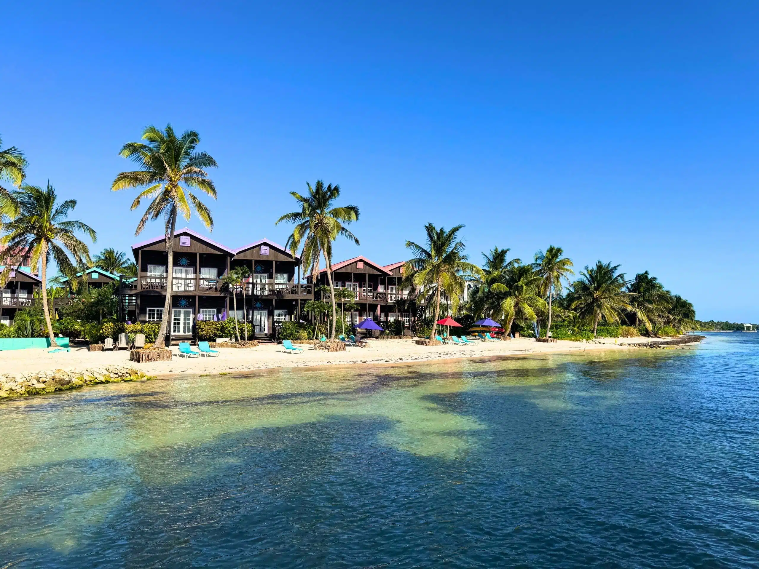 Panoramic waterfront view of Xtan Ha Resort on Ambergris Caye, Belize, from the calm turquoise Caribbean Sea, showing a row of dark timber two-storey beachfront villas with wrap-around balconies and colourful rooflines, teal sun loungers and purple and red parasols on the white sandy beach, lined with tall coconut palm trees under a vivid blue sky.