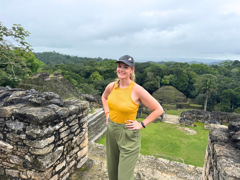 Smiling Michelle L. Jensen (Travel HerStory) in a yellow tank top, olive green trousers, and a dark baseball cap standing atop the Caana pyramid at Caracol, Chiquibil Forest Reserve, Belize, with hand on hip, overlooking moss-covered Maya temple structures, a green plaza, and an expansive Belizean jungle canopy stretching to the horizon.