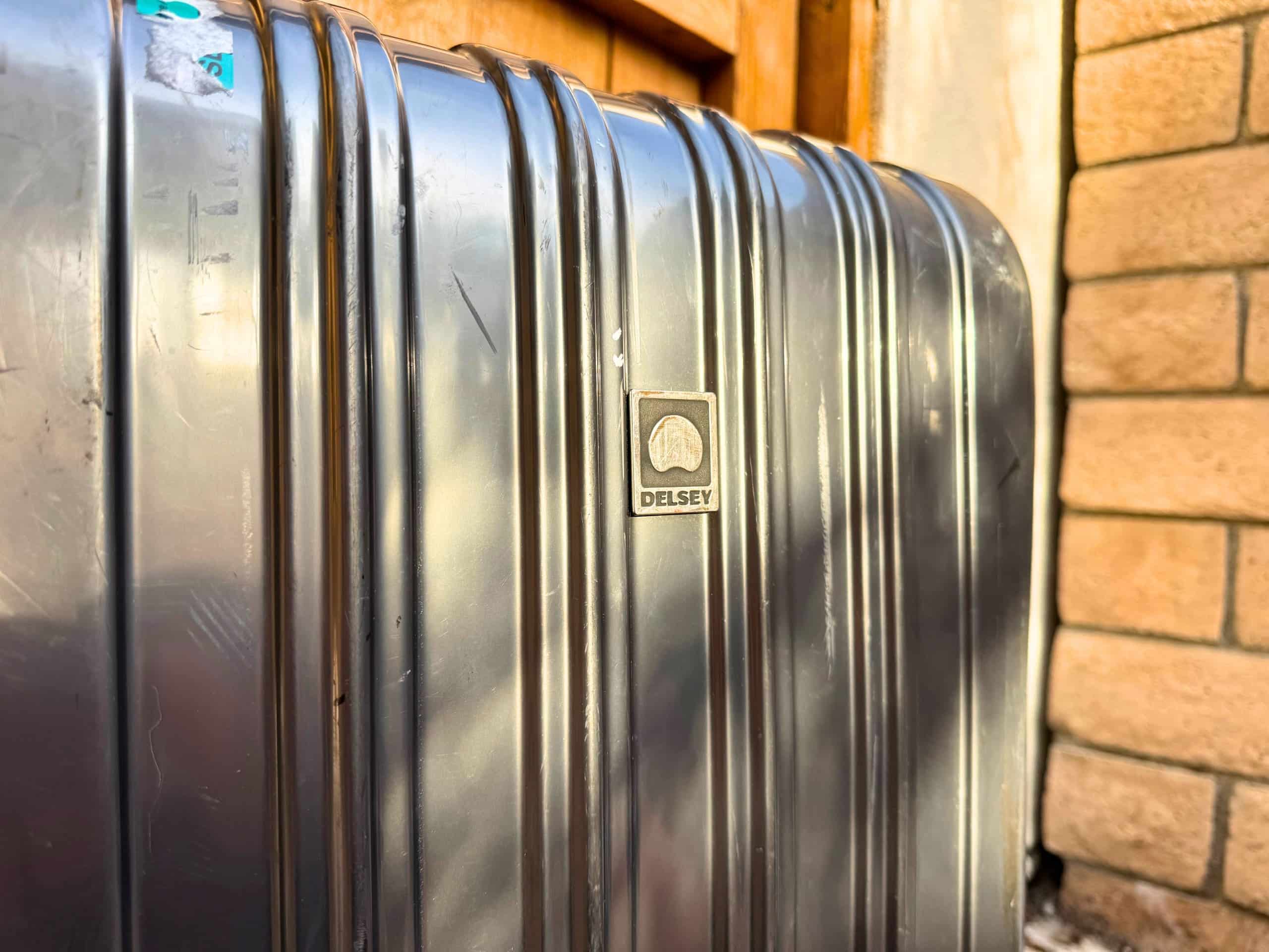 Close-up of the ribbed silver hardshell exterior of a Delsey Paris Helium Aero hardside suitcase showing the embossed Delsey logo badge at center, with minor surface scratches visible on the polished grooved shell, propped against a brick wall.