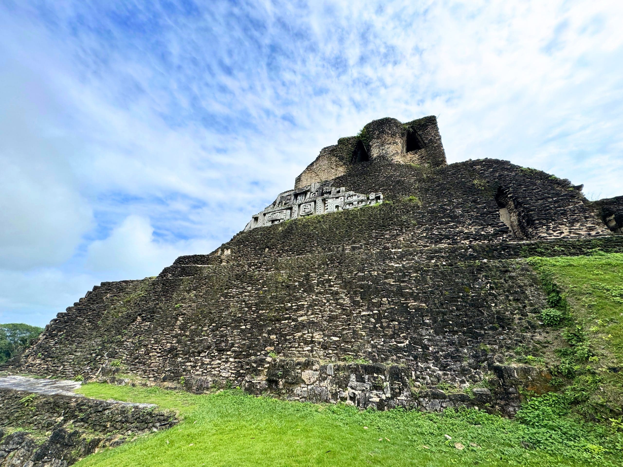 Home 29 Low-angle view looking up at El Castillo, the imposing dark stone pyramid at Xunantunich Maya ruins near San Ignacio Belize. Intricate carved stone friezes depicting Maya deities are visible near the partially ruined summit. Lush green grass covers the foreground terrace and vegetation edges the stepped stone walls under a bright blue sky with wispy white clouds.