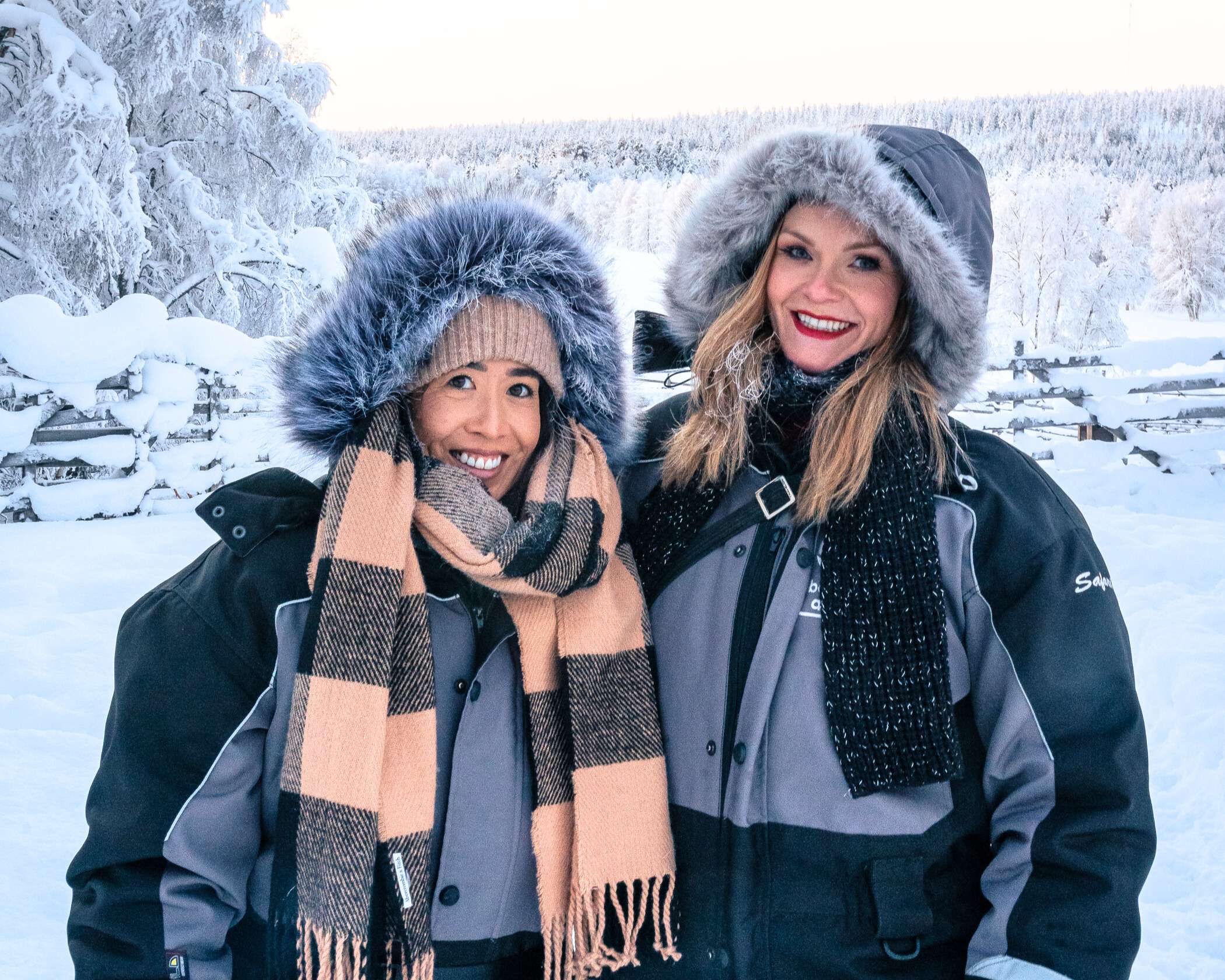 Michelle L. Jensen (Travel HerStory) and a friend posing in a snowy Finnish landscape in snow suits and scarves before a snowy Lapland forest.