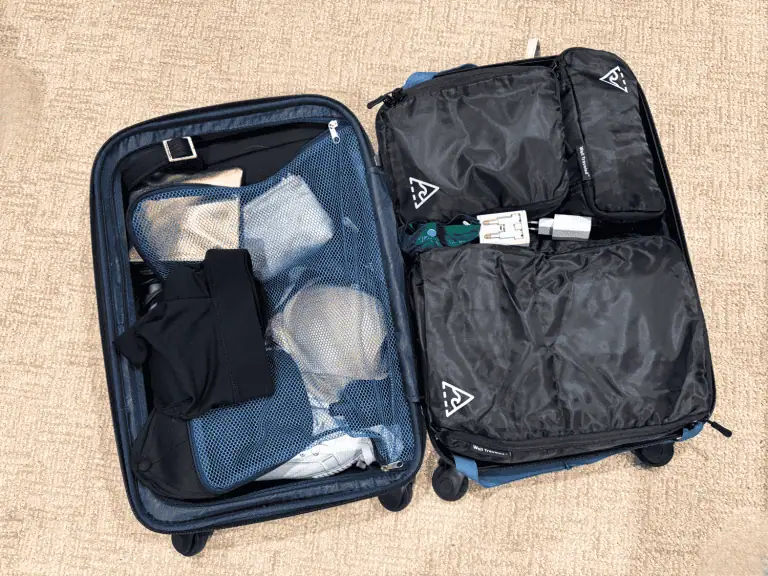 Open blue hardside suitcase on a carpet showing three black Well Traveled packing cubes neatly organized in the main compartment alongside a travel adapter and accessories, with the opposite side featuring a blue mesh panel holding shoes and undergarments.