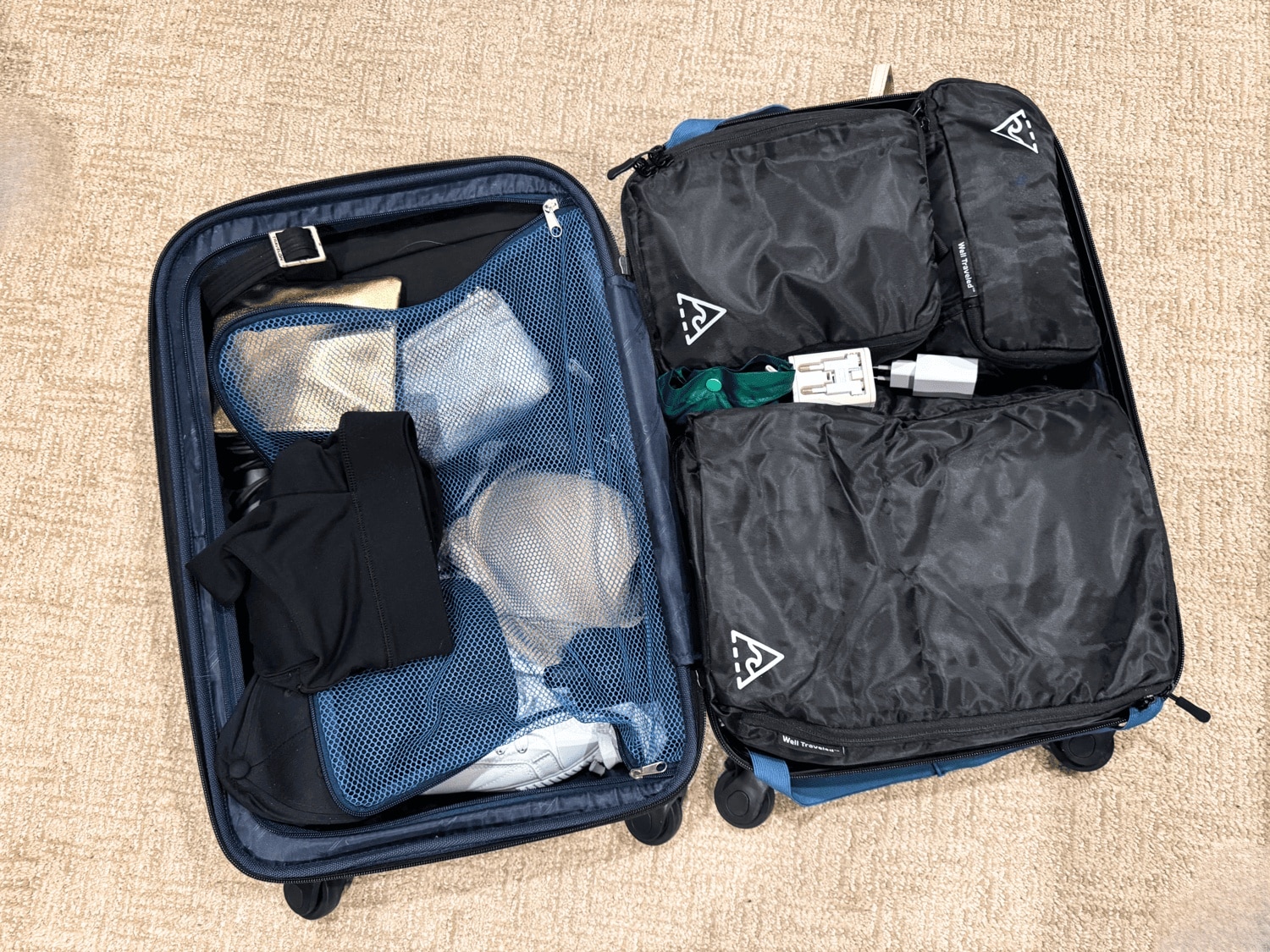 Open blue hardside suitcase on a carpet showing three black Well Traveled packing cubes neatly organized in the main compartment alongside a travel adapter and accessories, with the opposite side featuring a blue mesh panel holding shoes and undergarments.