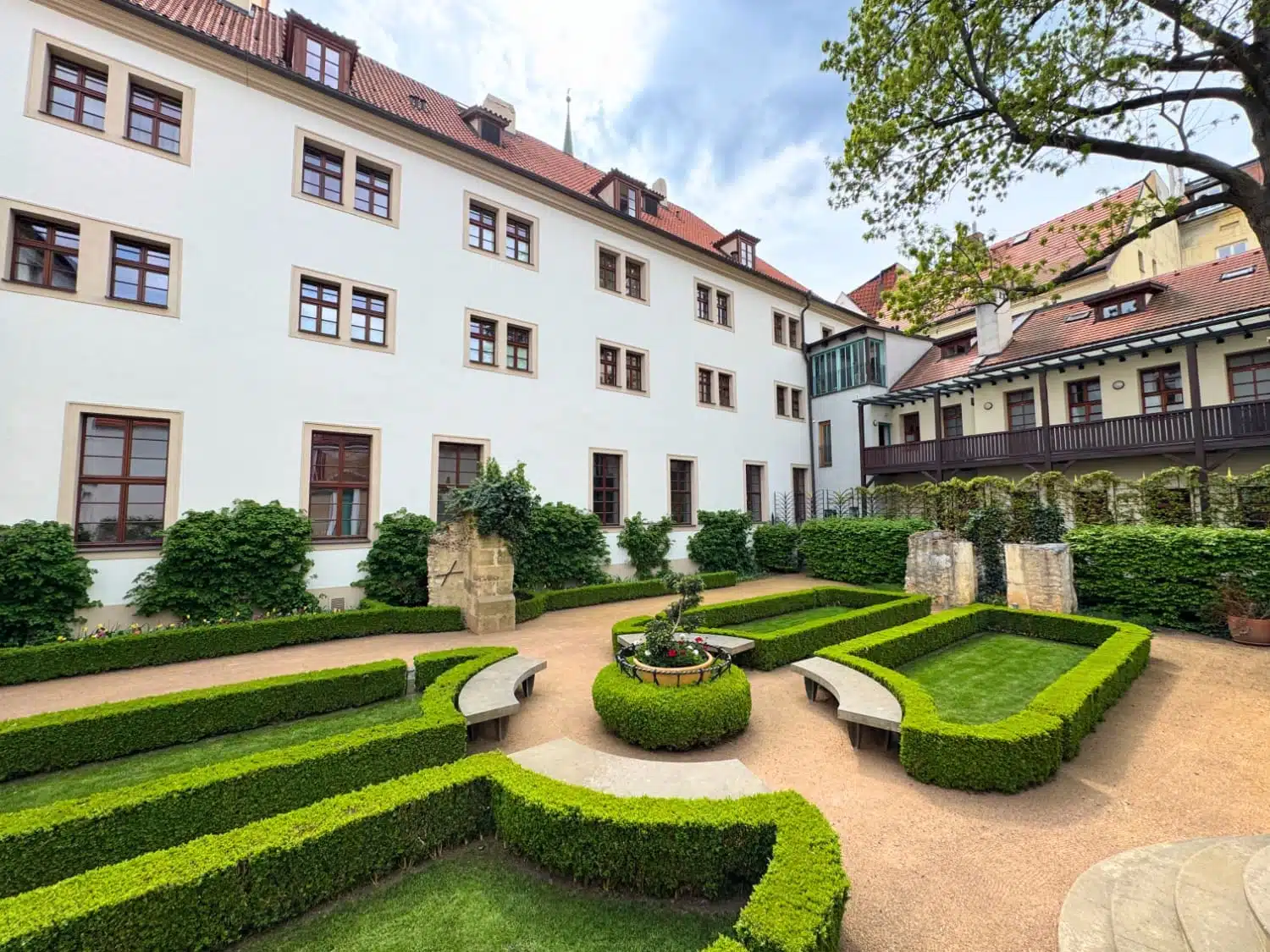 Formal Baroque courtyard garden at Augustine, a Luxury Collection Hotel in Prague, Czech Republic, featuring manicured boxwood hedge parterres, curved stone benches, a central flower planter, and the hotel's historic white monastery building framing the peaceful space.