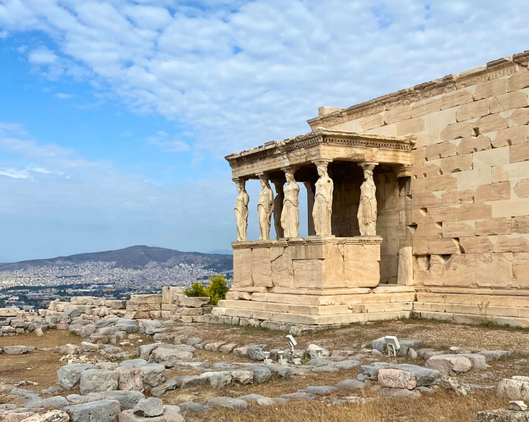 Six sculpted female figures known as Caryatids support the porch roof of the Erechtheion, an ancient Greek temple on the Acropolis of Athens, Greece. The marble statues stand in graceful poses, serving both decorative and structural roles against a backdrop of clear blue sky.