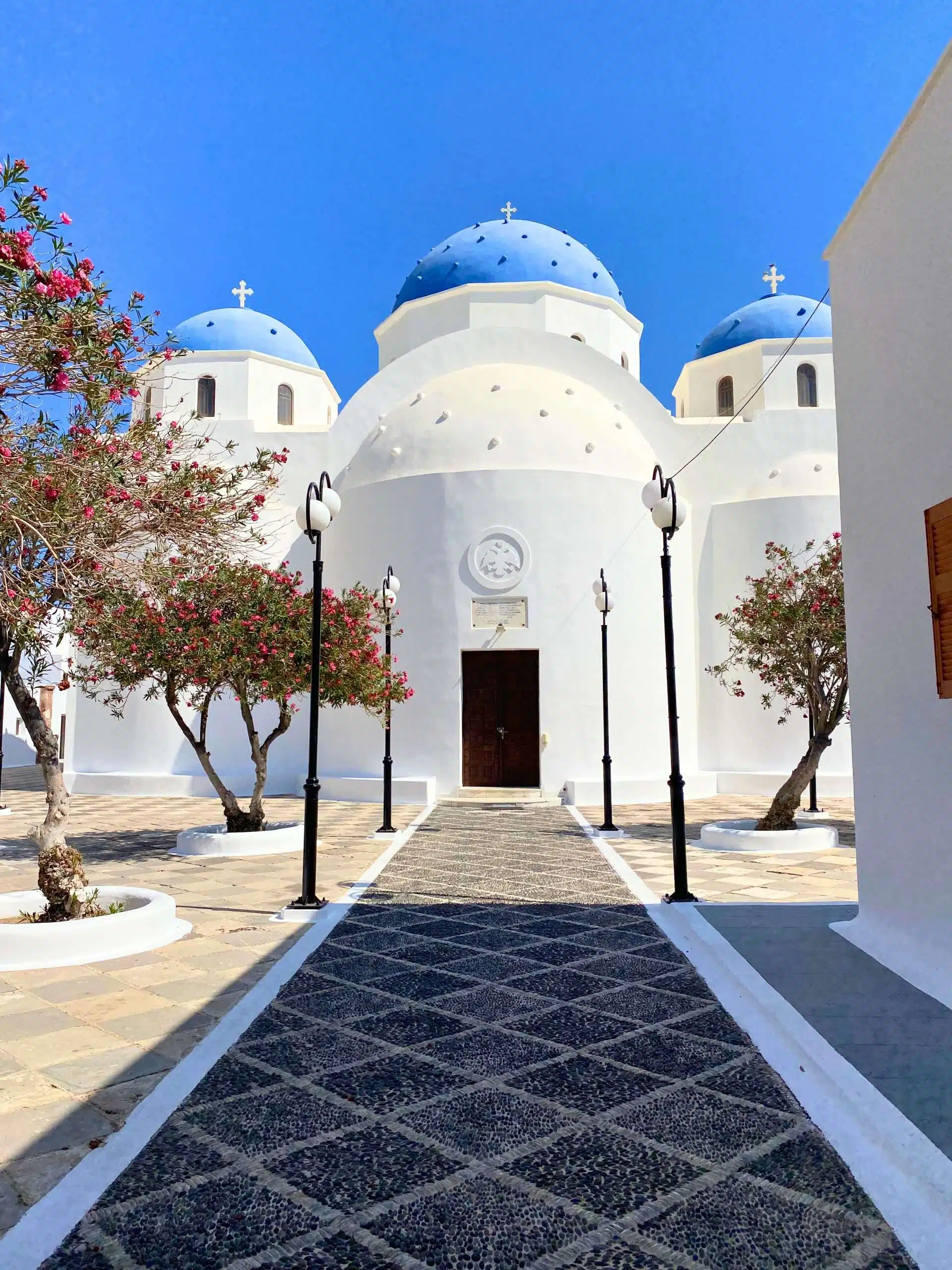 Home 31 The Church of the Holy Cross in Santorini, Greece, featuring a large white facade and a bright blue dome topped with a cross. The church stands out against a clear sky, as a stone path leads to it with pink flowers blooming to the sides.