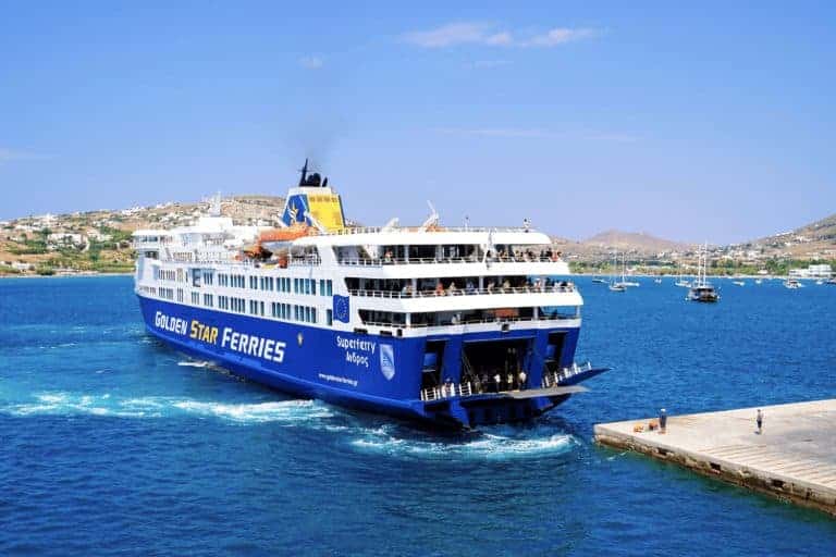 Travelers on the deck of a Golden Star Ferry with Greek islands in the distance and clear blue skies overhead, the vessel’s bold blue and yellow branding clearly visible against the calm sea as the vessel docks.