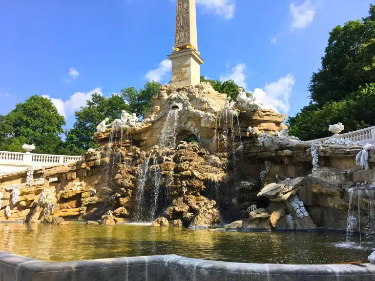 The Obelisk Fountain at Schonbrunn Palace, featuring cascading water, sculpted figures, and a tall obelisk rising from a rocky base surrounded by lush greenery under a blue sky in Vienna, Austria.