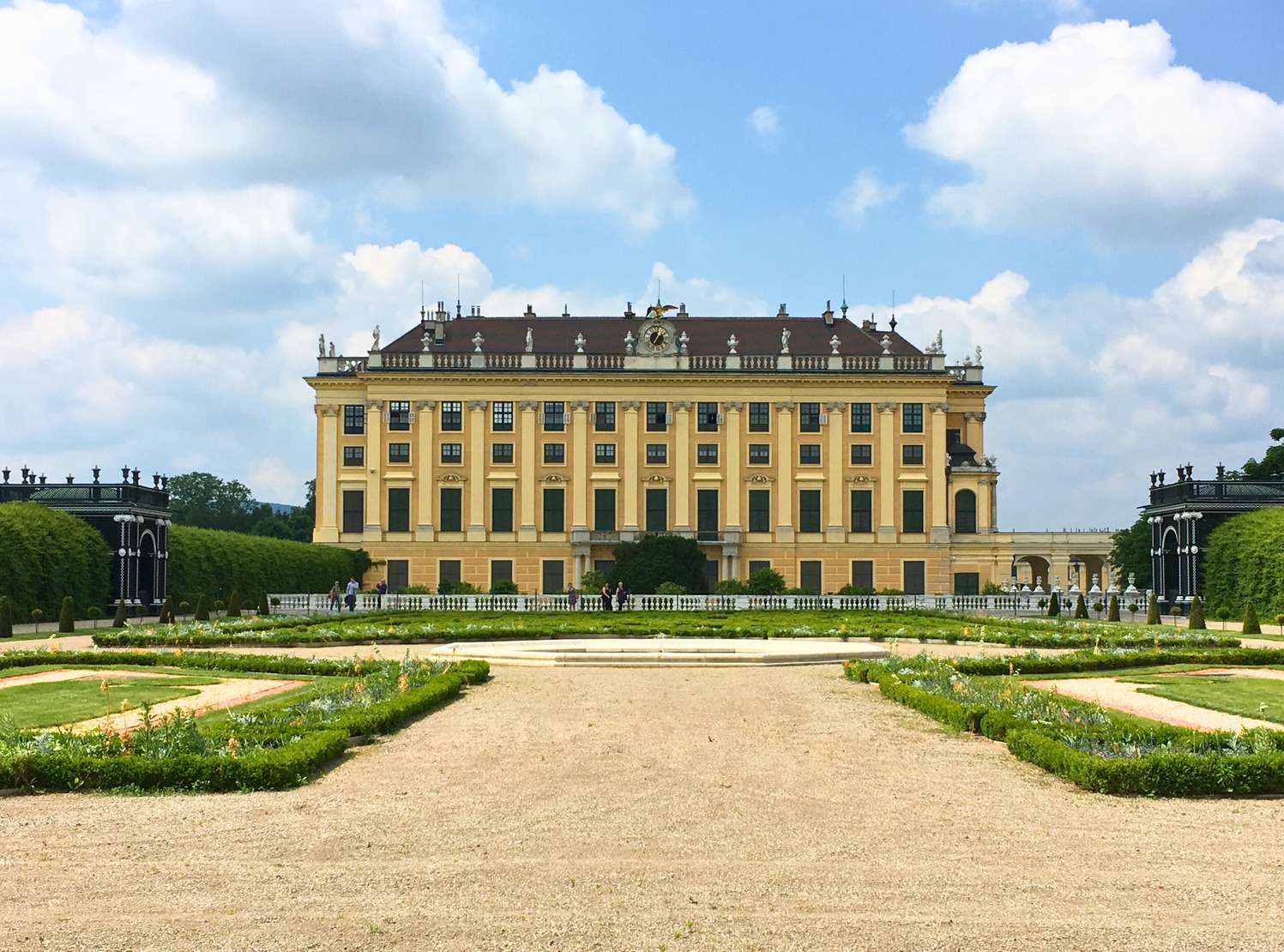 The rear exterior of Schonbrunn Palace with its symmetrical yellow façade and clock overlooking manicured gardens and pathways under a blue sky in Vienna, Austria.