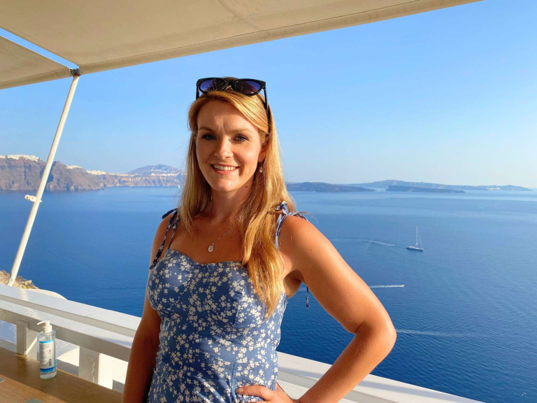 Michelle L. Jensen (Travel HerStory) in a breezy blue floral dress smiling before a coastal Greek view of the Aegean from the Pelekanos Restaurant in Oia, Santorini, Greece.