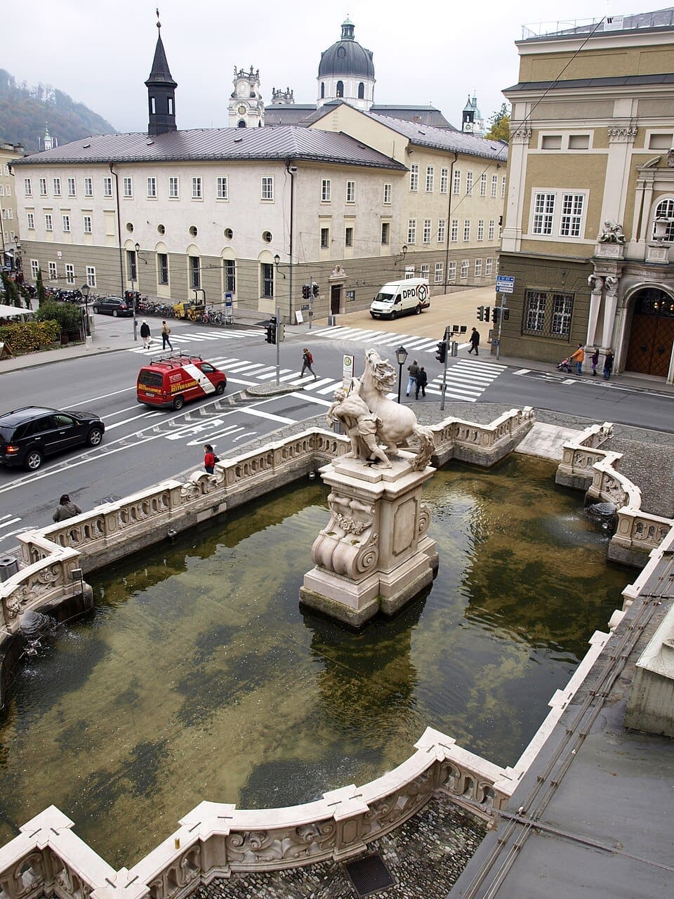 13 Sound of Music Filming Locations in Salzburg, Austria + How to Visit Them 43 Aerial view of the Baroque Pferdeschwemme horse pond fountain on Karajan-Platz in Salzburg, Austria, featuring a rearing horse sculpture, with the domes and towers of Kollegienkirche visible in the background beyond a busy street intersection.