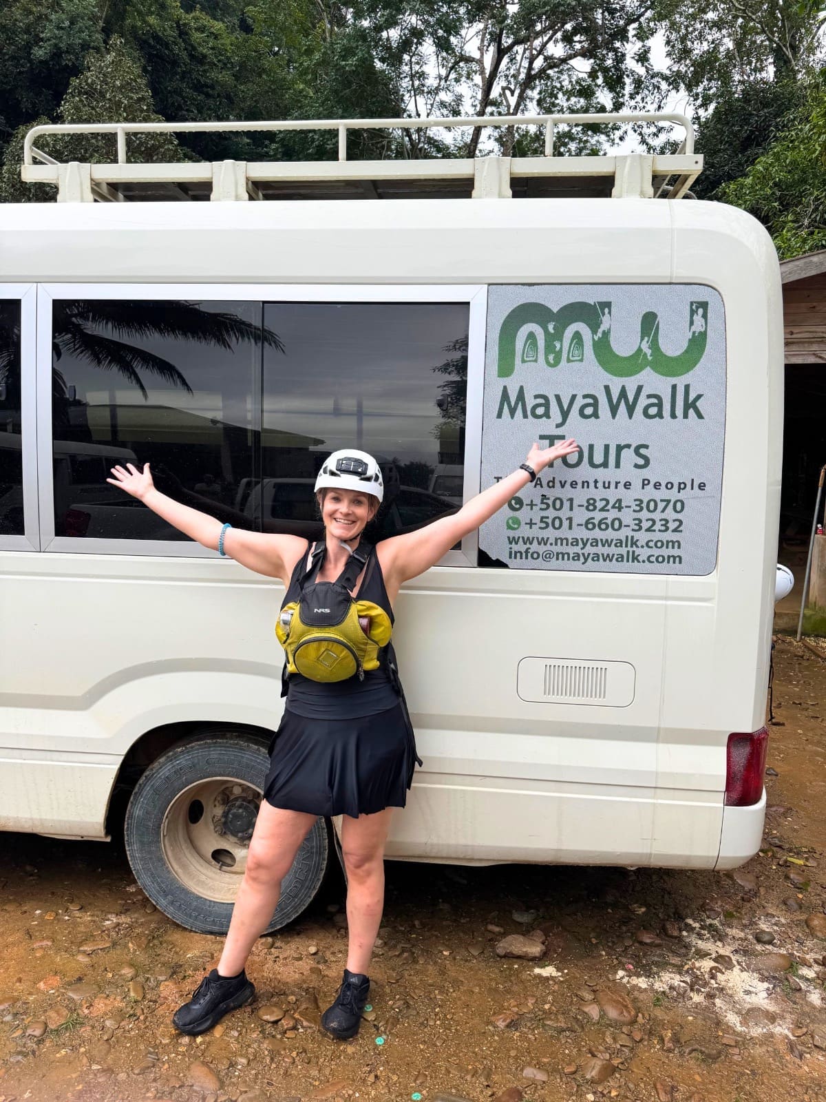My Experience Staying in a Hostel as a Solo Female Over 35: Honest Insights 9 A woman wearing a bathing suit, a hard hat with a headlamp, and a life vest stands before a white tour bus with "MayaWalk Tours" branding parked on a dirt path surrounded by lush green jungle foliage. The bus is preparing for the Actun Tunichil Muknal (ATM) cave tour in Belize.