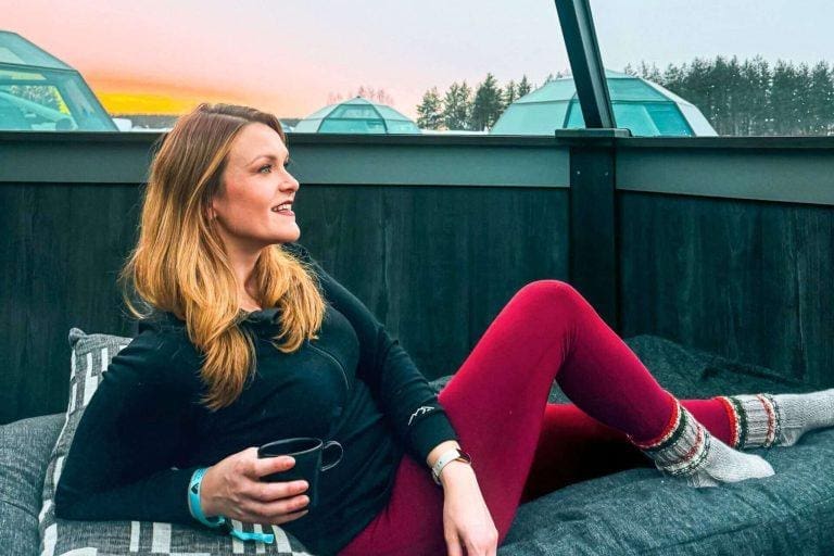 Michelle L. Jensen (Travel HerStory) lounging in a glass igloo hotel room at the Arctic SnowHotel & Glass Igloos in Sinettä, Finland, bundled in winter clothing with snow-covered trees and a soft pink sky in the background.