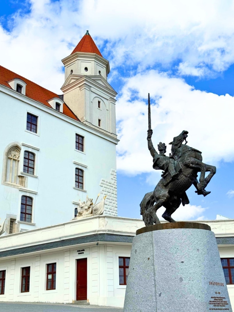 Destinations 15 Bronze equestrian statue of Svätopluk I on a rearing horse with sword raised, mounted on a granite pedestal in the courtyard of Bratislava Castle, with the white castle facade featuring a red-roofed corner tower and a decorative stone relief visible behind it under a blue sky in Slovakia.