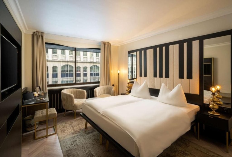 Where to Stay in Vienna, Austria: My Favorite Areas + Best Budget Hotels 8 Guest room at Elaya Hotel Vienna City Center in Vienna, Austria, featuring a piano-key patterned headboard, wall-mounted TV, gold accent lamps, and a city street view through the window.