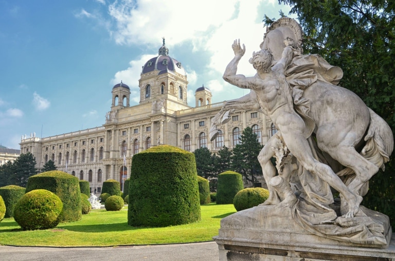 Exterior of the Kunsthistorisches Museum in Vienna, Austria, with its grand dome, ornate stone facade, statue, and symmetrical landscaping leading up to the main entrance under a blue sky.