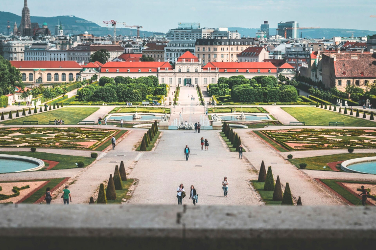 Baroque Lower Belvedere Palace with its ornate white facade, orange colored roofs, and symmetrical gardens dotted with ponds under a clear sky with the city of Vienna, Austria, in the background.